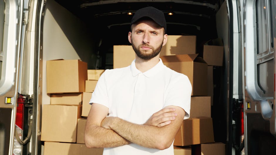 A young man with a serious expression, wearing a white polo shirt and a black cap, stands with his arms crossed in front of an open commercial van loaded with numerous packed cardboard boxes and some 