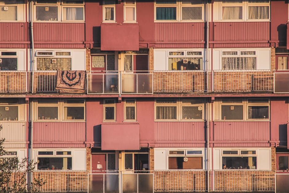 A multi-storey residential building with a pink exterior, featuring several balconies enclosed by metal and glass railing panels, some of which have curtains or fabric coverings visible through the wi