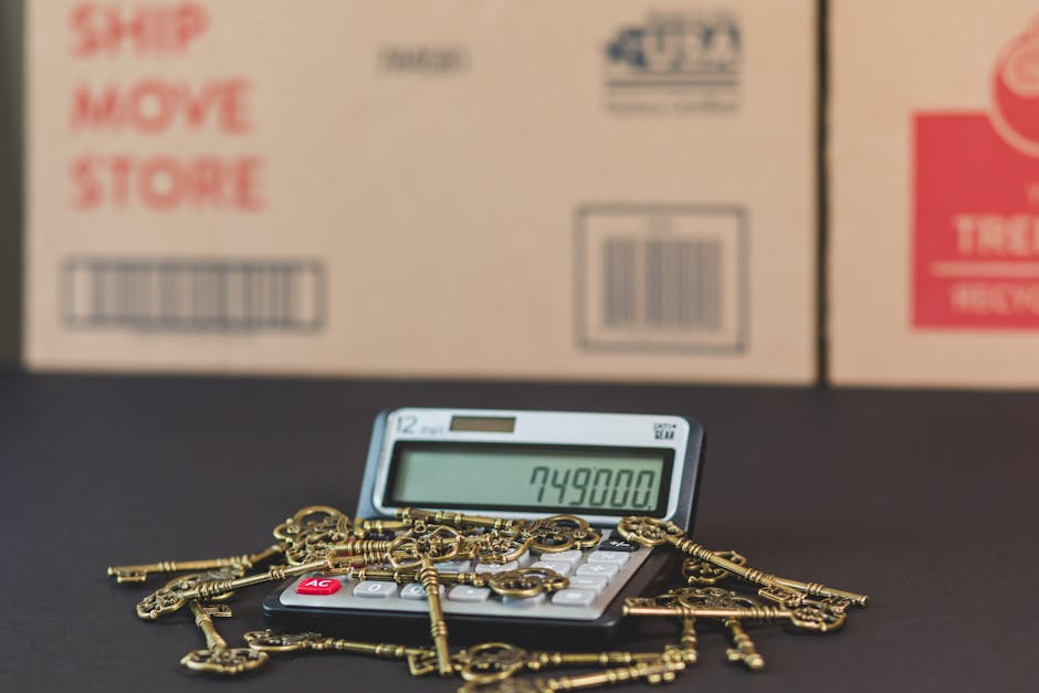 A digital calculator displaying the number 749800 is positioned on a black surface, with numerous antique brass keys and keychains scattered around it. In the background, blurred cardboard boxes marke
